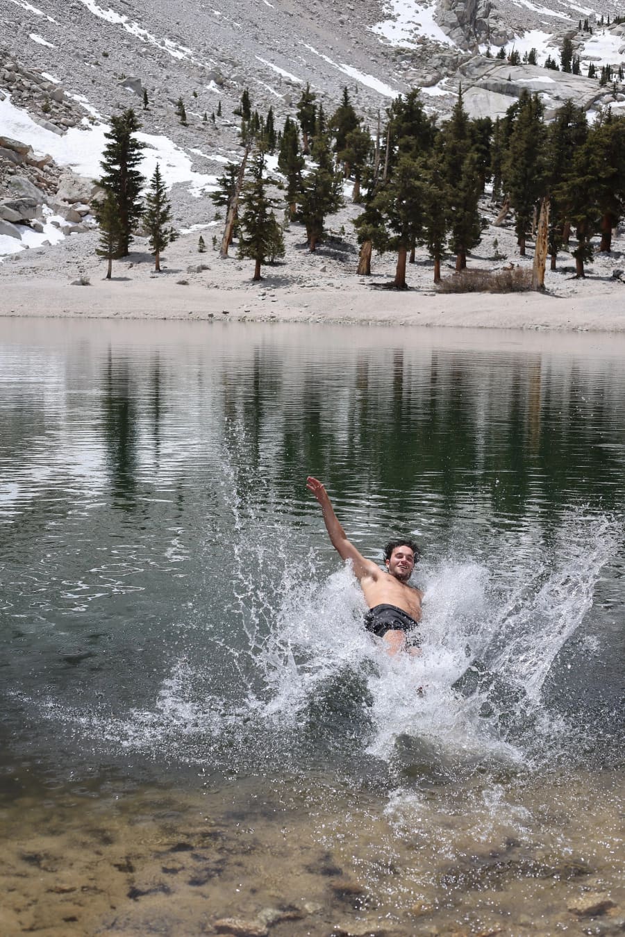 A swimmer mid-air in a wild plunge into a glacial alpine lake, ringed by snow patches and small evergreens.