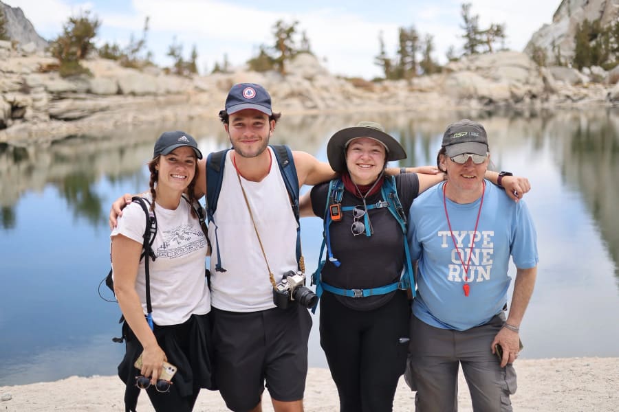 Four hikers in caps and packs huddle together for a smiling group photo by the still water of a high-mountain lake.