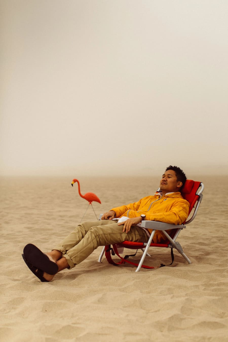 A man in a yellow windbreaker reclines in a low beach chair on hazy sand beside a small plastic flamingo.