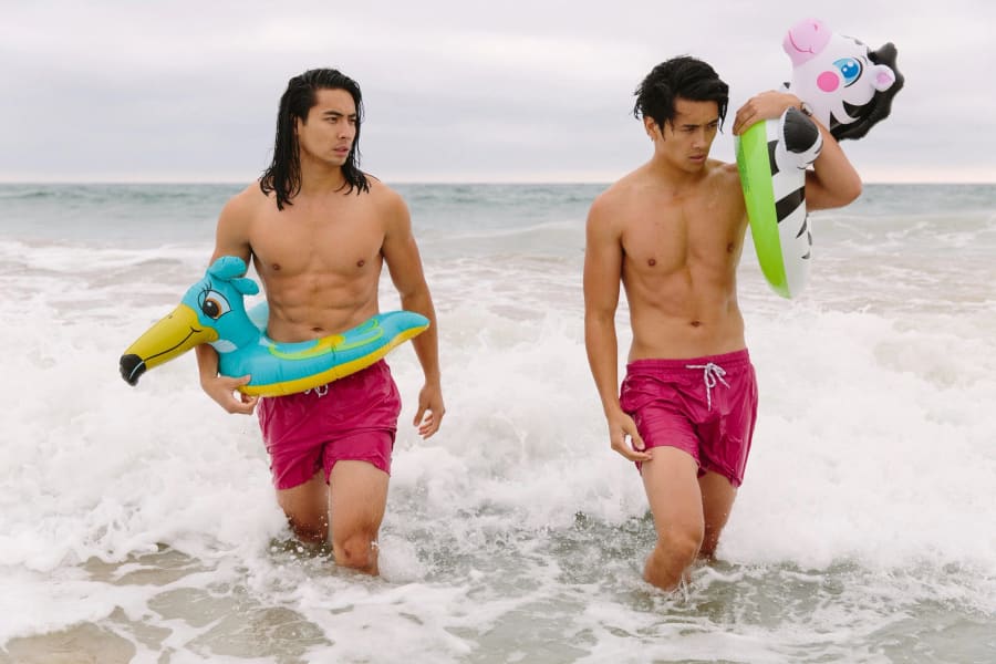 Two men in pink swim trunks wade out of breaking surf, carrying inflatable pool floats shaped like a flamingo and a zebra.