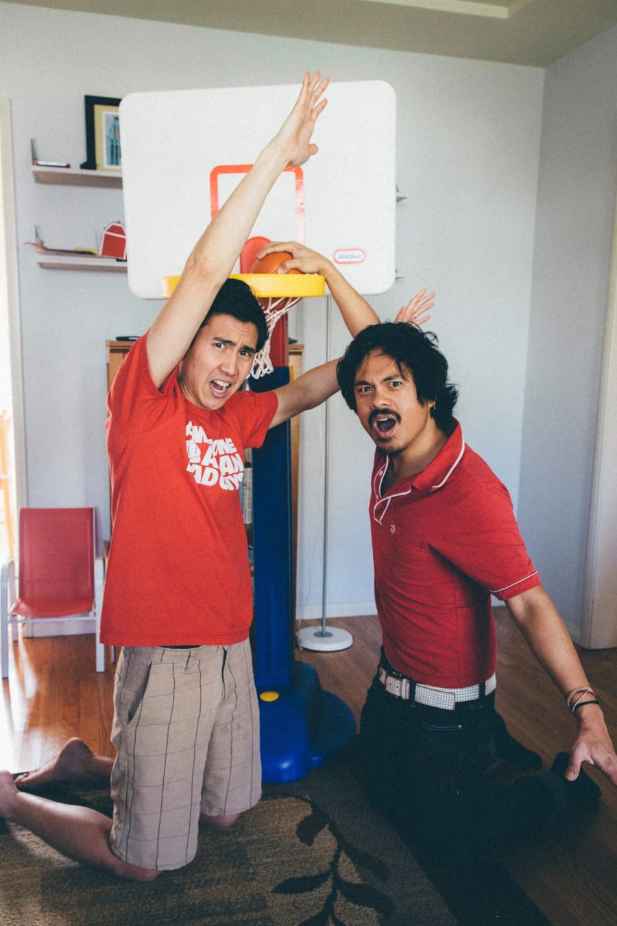 Two men in red shirts kneel beside a toy basketball hoop, arms raised mid-celebration in a sunlit living room.