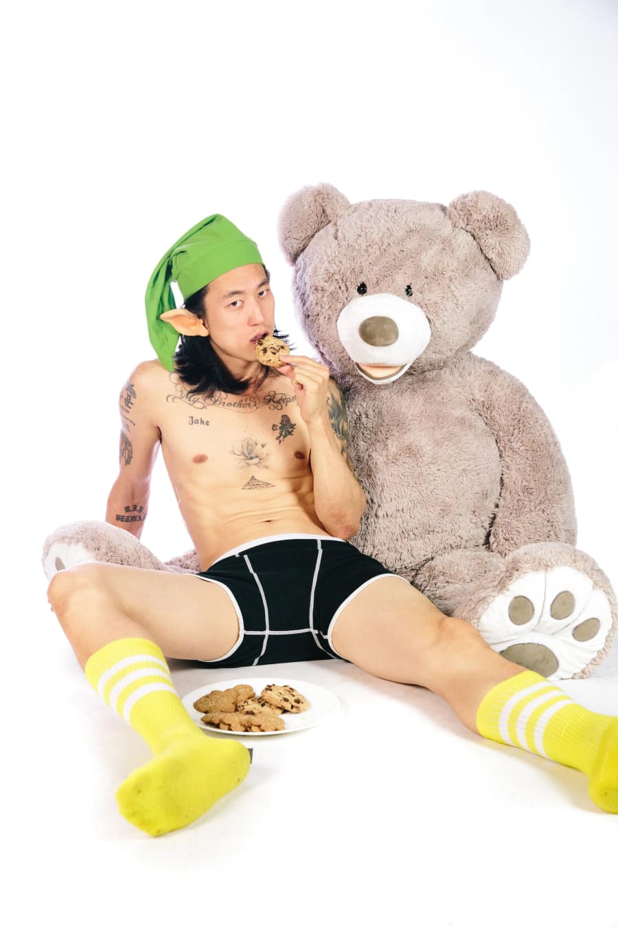 A man wearing only an elf hat, briefs and yellow socks sits beside a giant teddy bear, eating a chocolate-chip cookie.