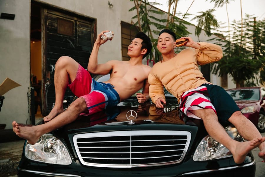 Two shirtless men in swim trunks lounge across the hood of a black Mercedes on a side street, holding burgers up to the light.