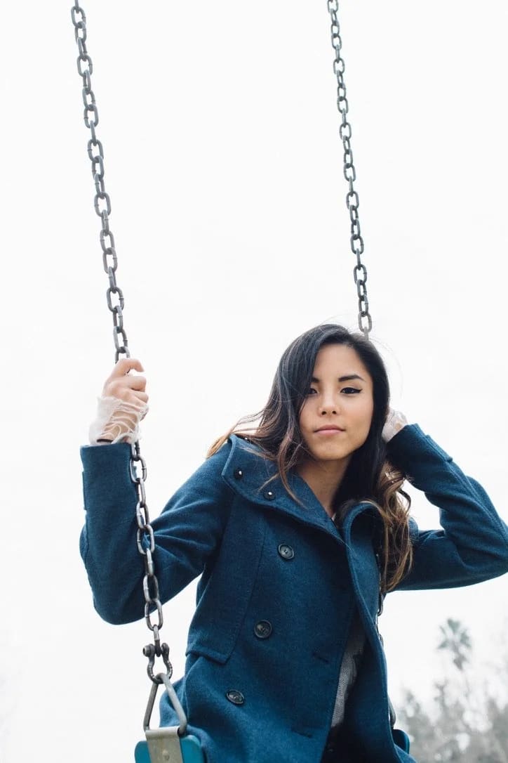 A young woman in a navy peacoat sits on a playground swing, hand resting against her hair under a soft overcast sky.