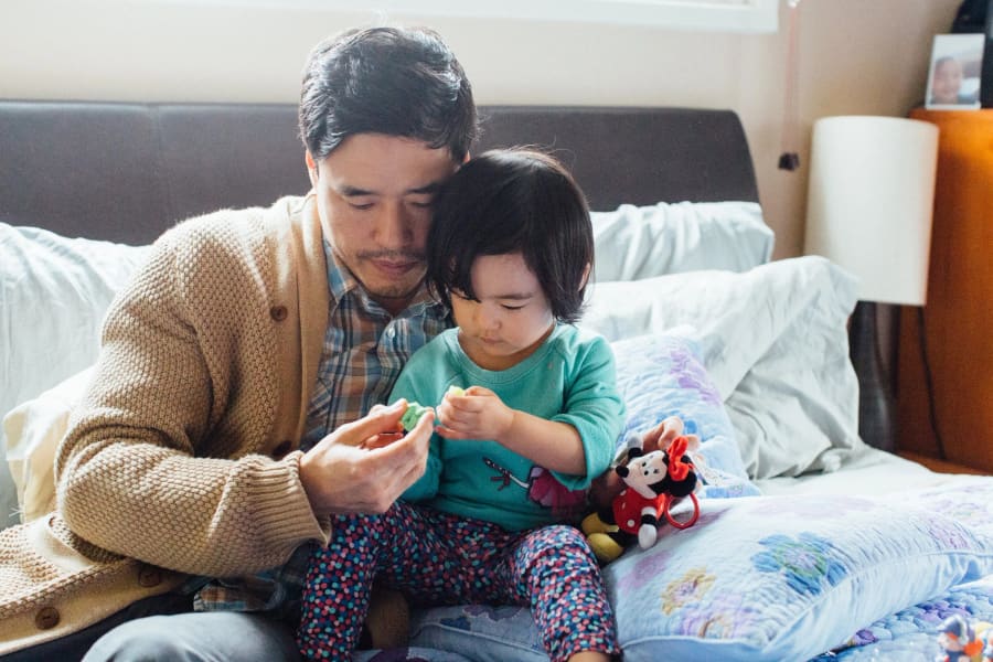 A father in a tan cardigan and his small daughter share a quiet moment on a couch, examining a toy together.