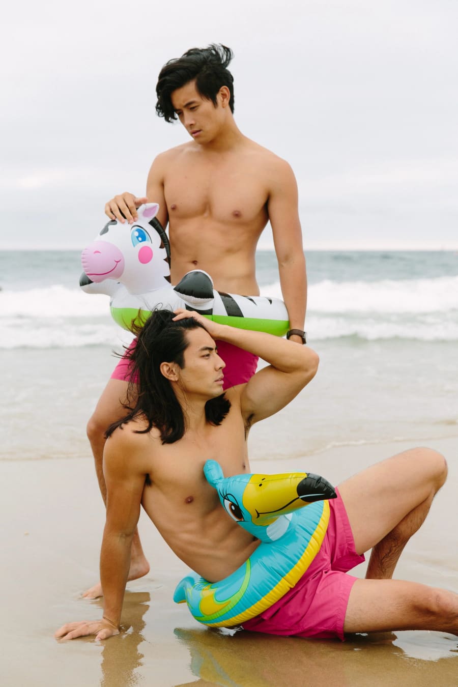 Two shirtless men in pink shorts pose on a wet beach with cartoon-themed inflatable pool floats, ocean breaking behind them.