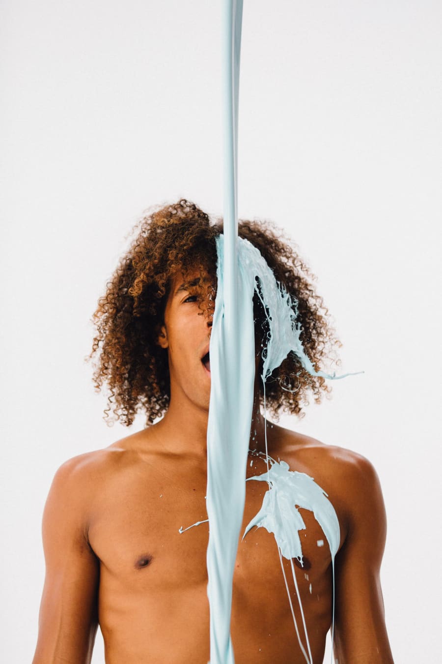 A shirtless young man with curly hair stands as a stream of pale blue paint pours down across his face and chest.