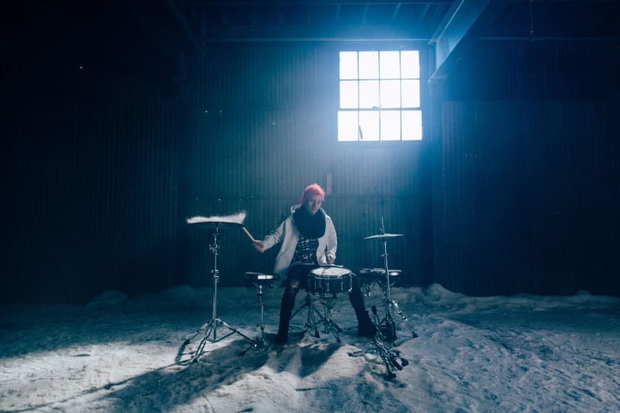 A drummer with bright red hair plays a kit in an industrial warehouse, snow falling around them under a single window beam.