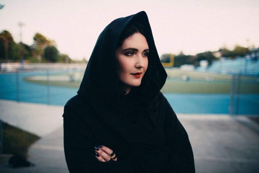 A woman in a black hooded cloak with red lipstick stands on a school running track at dusk, arms folded.