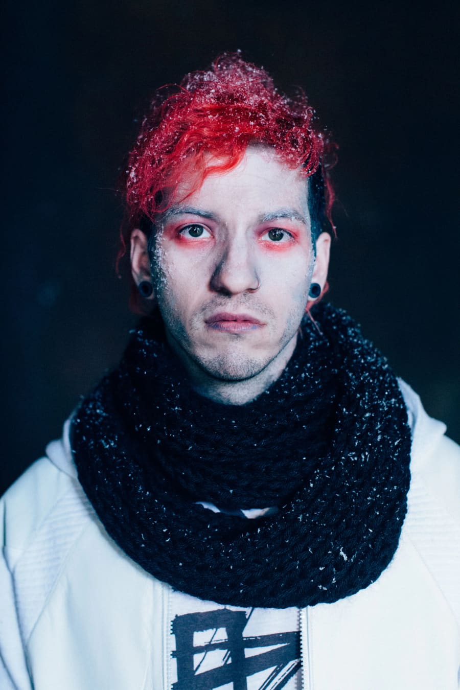 Tight portrait of a young man with bright red curly hair and red-rimmed eye makeup, wrapped in a chunky black scarf.
