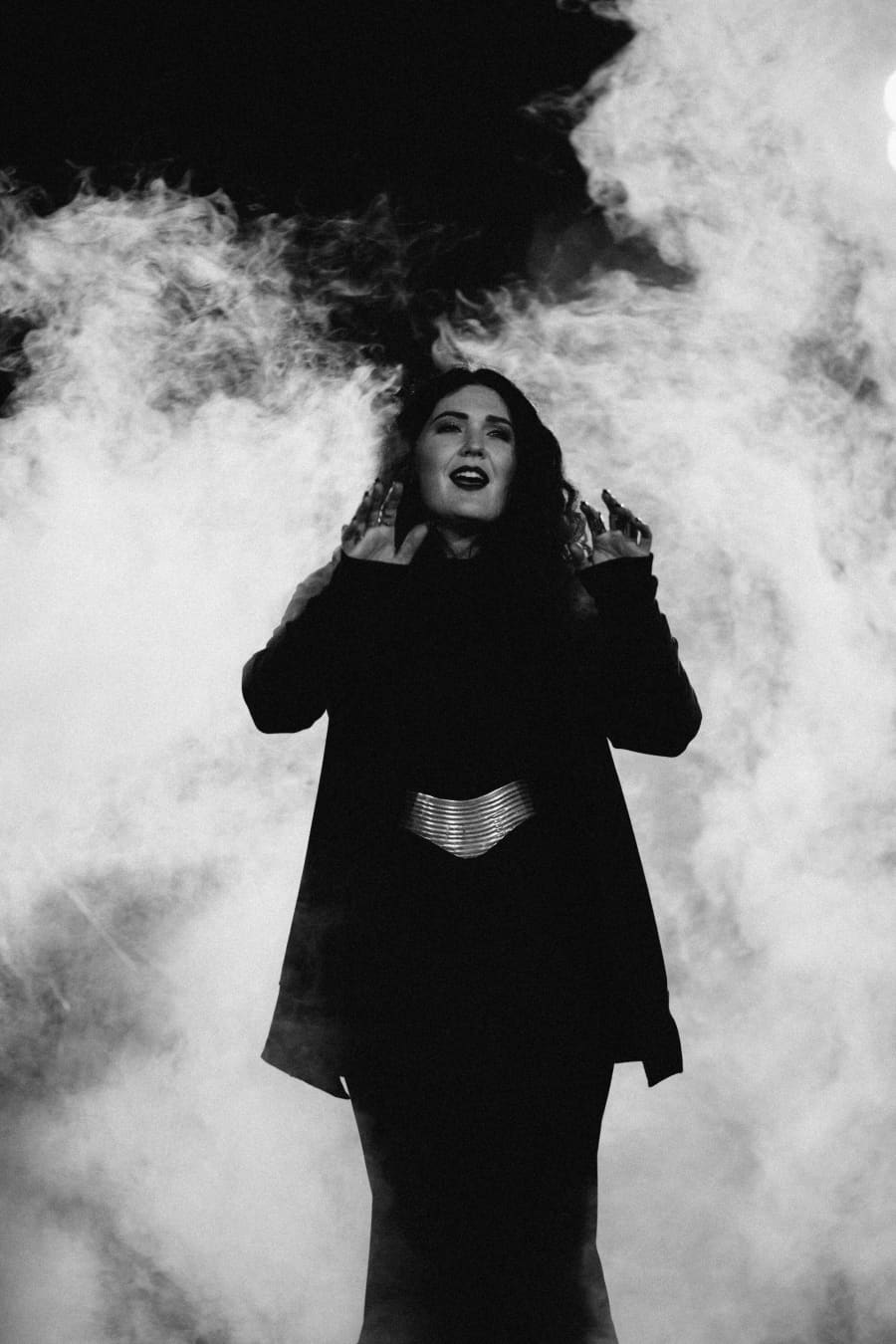 Black-and-white image of a woman in a long black coat throwing up middle fingers, swirling smoke filling the frame.
