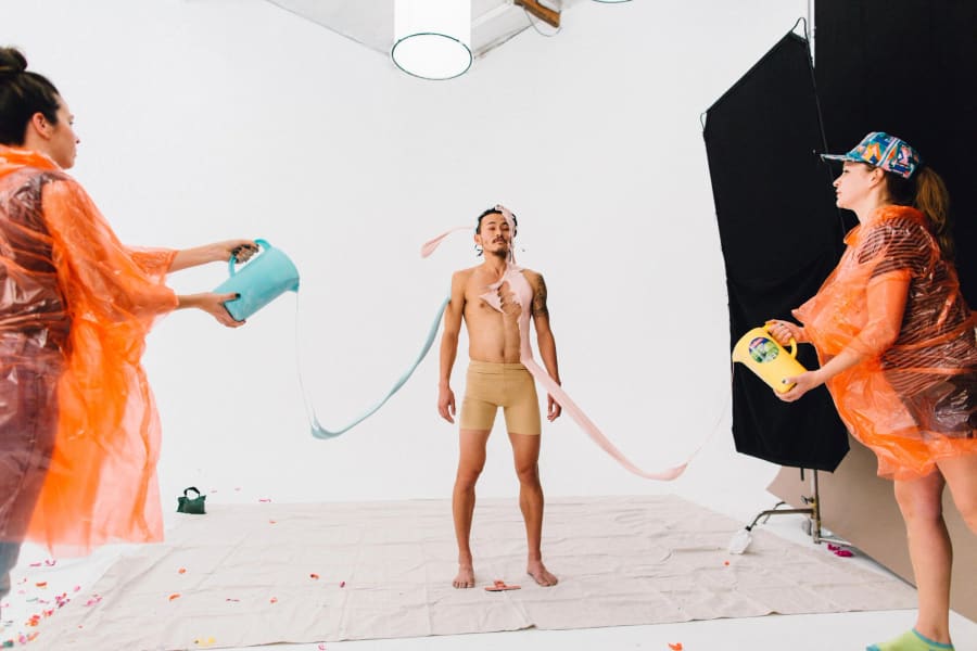 Two crew members in bright orange rain ponchos pour buckets of liquid over a model in tan briefs against a black backdrop.
