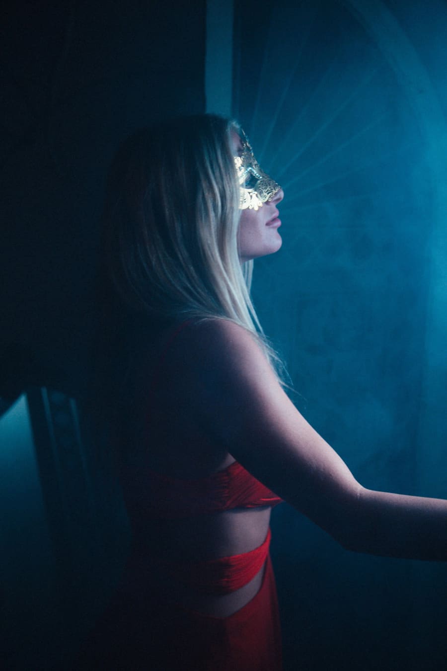 A blonde in red lingerie and a delicate gold filigree mask stands in profile against a smoky blue-lit interior.