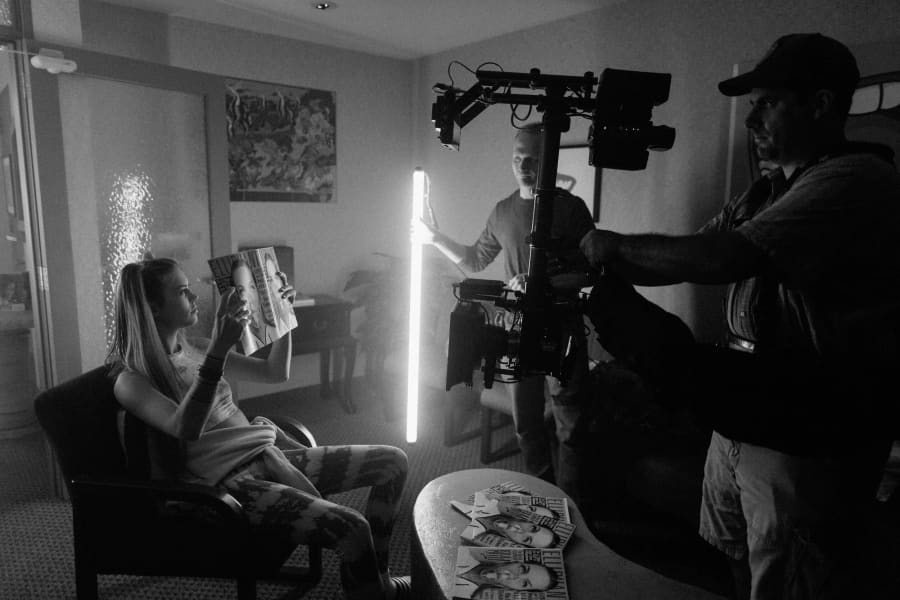 Behind-the-scenes black-and-white: an actress in a hotel chair holds a magazine while crew members operate a dolly with a long tube light.