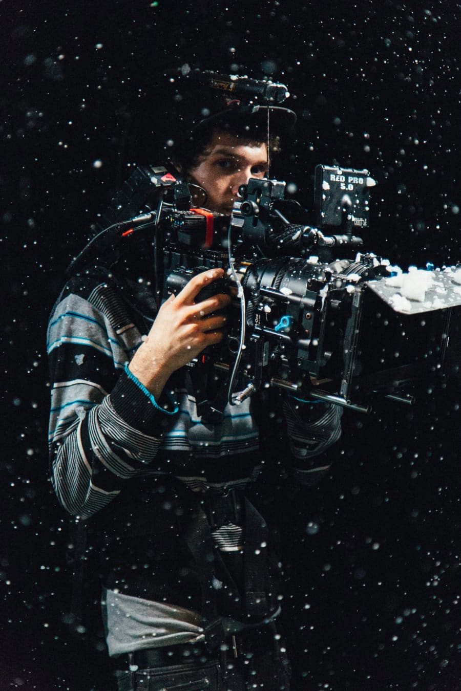 A cinematographer hand-holds a RED Pro 5.0 camera in a snow-flecked nighttime shot, framed in a vertical composition.